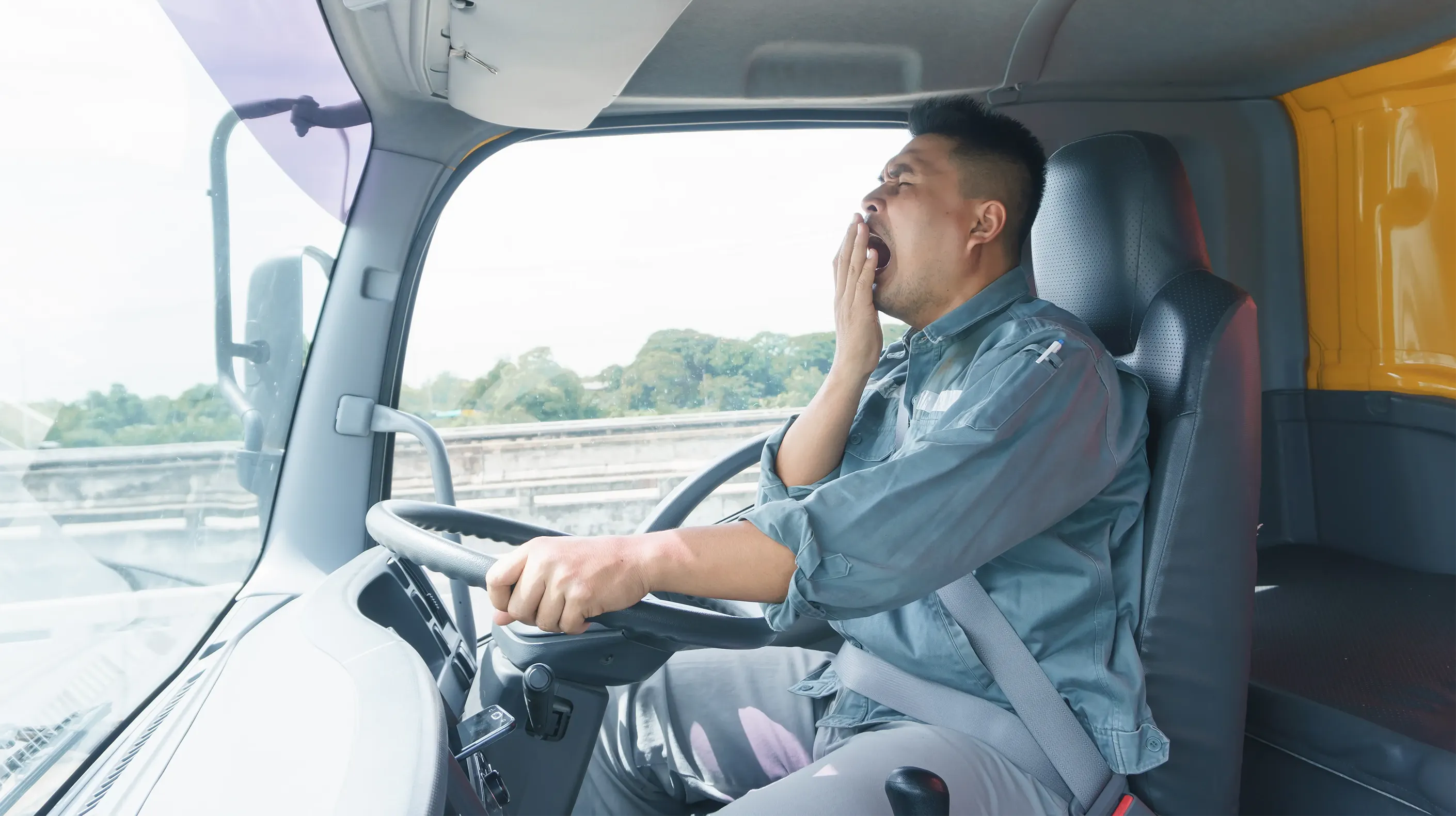 A tired truck driver yawning while holding onto the steering wheel with one hand.