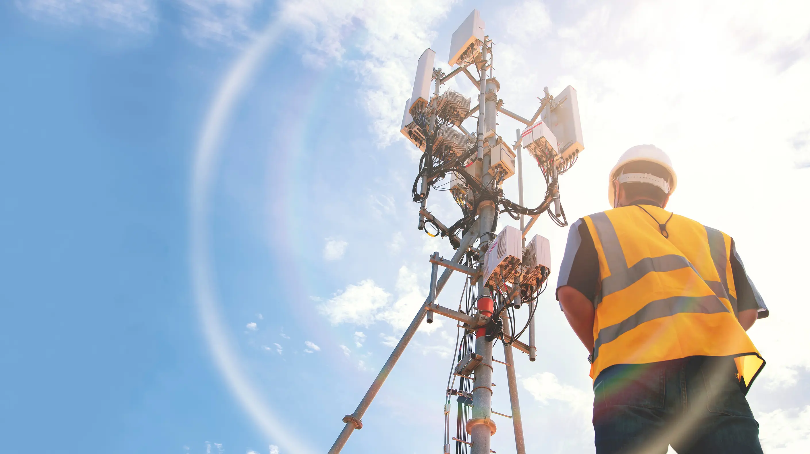 A worker wearing a safety vest staring up at a 5G tower.